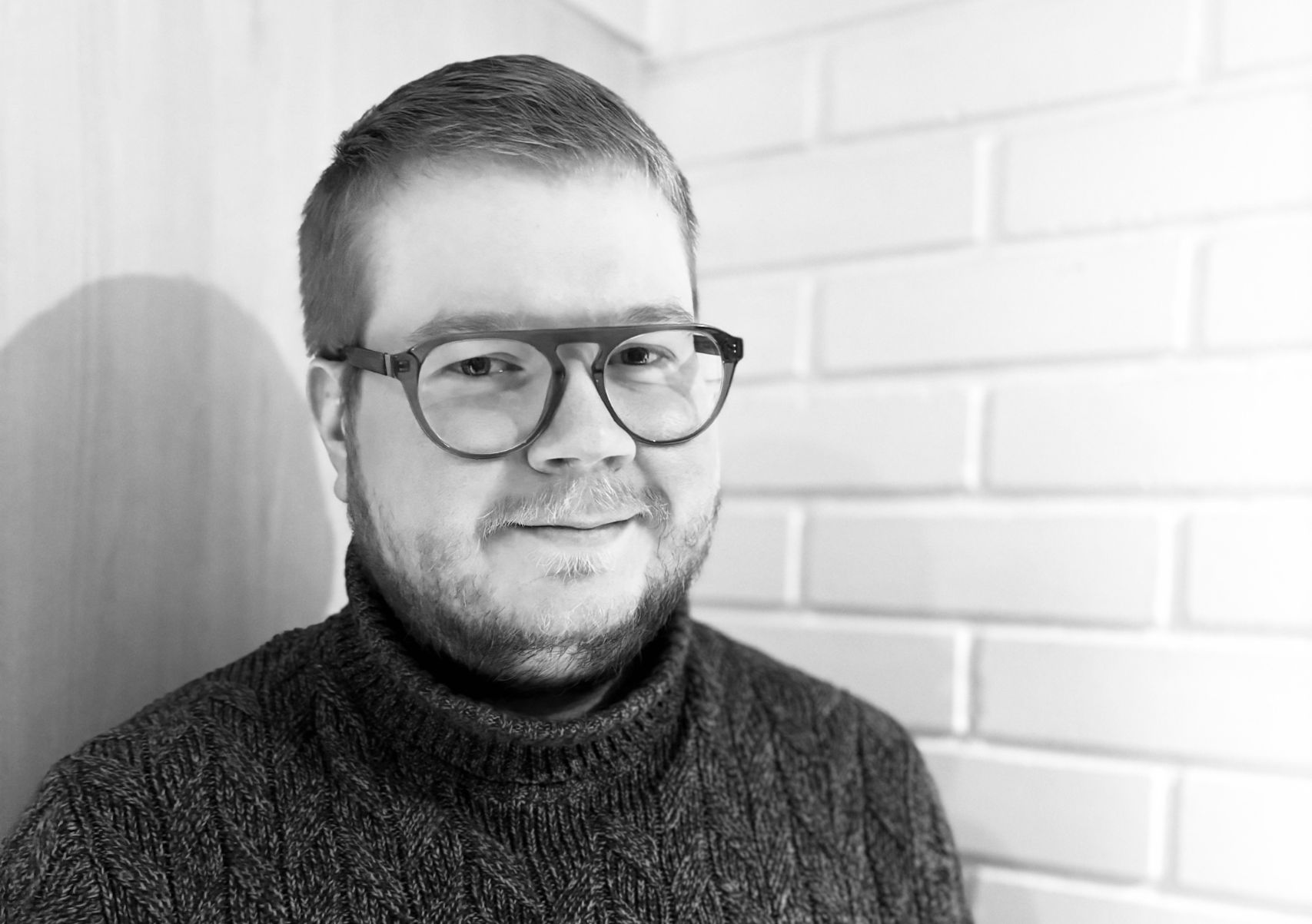 silmälasipäinen mies seisoo valkean tiiliseinän edessä