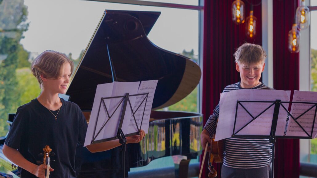 kaksi lasta seisovat nuottitelineiden takana viulut käsissään ja hymyilevät.