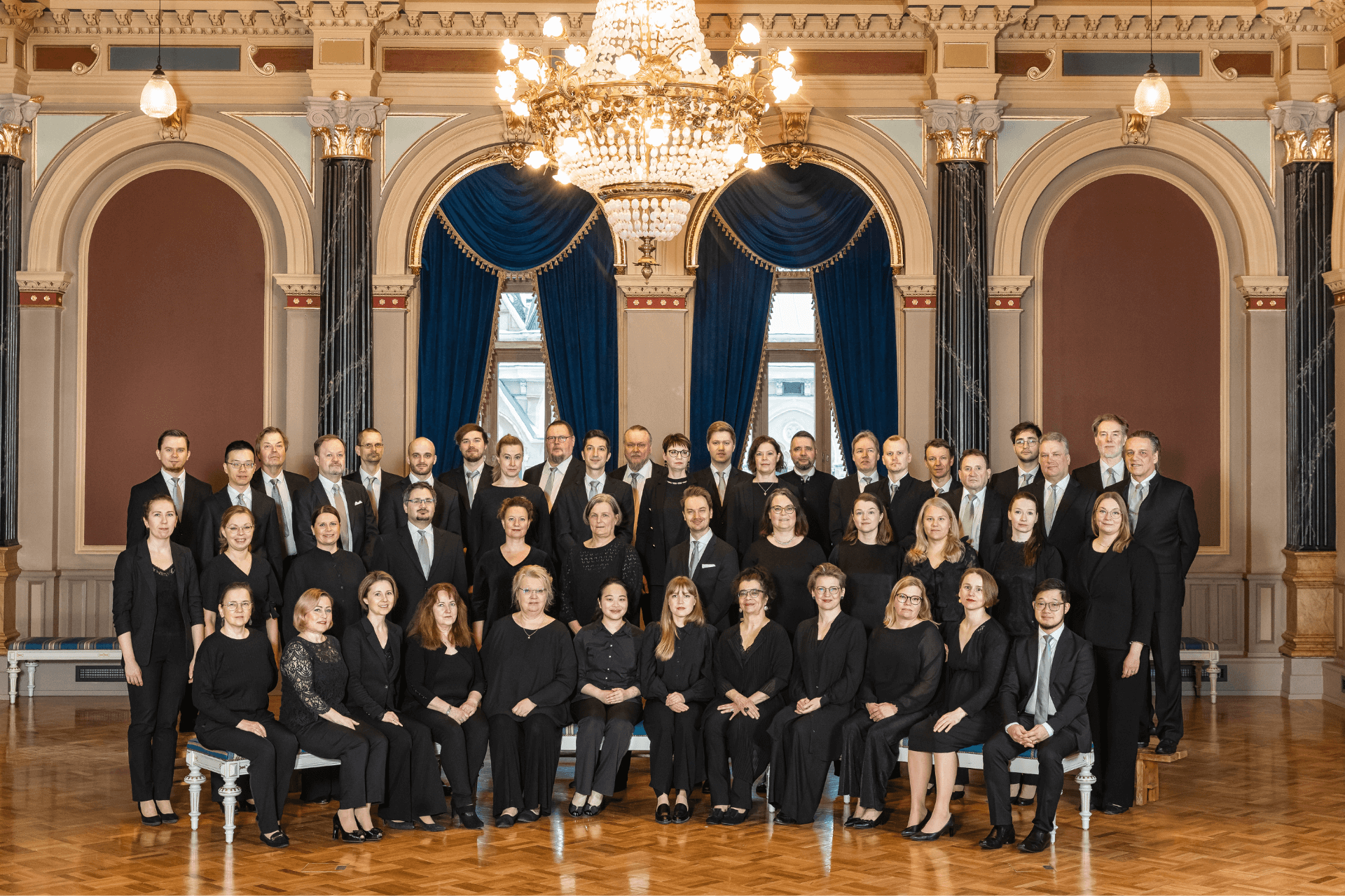 Iso ryhmä mustiin pukeutuneita ihmisiä istuvat ja seisovat koristeellisessa salissa. Kaikki katsovat kamarena suuntaan.