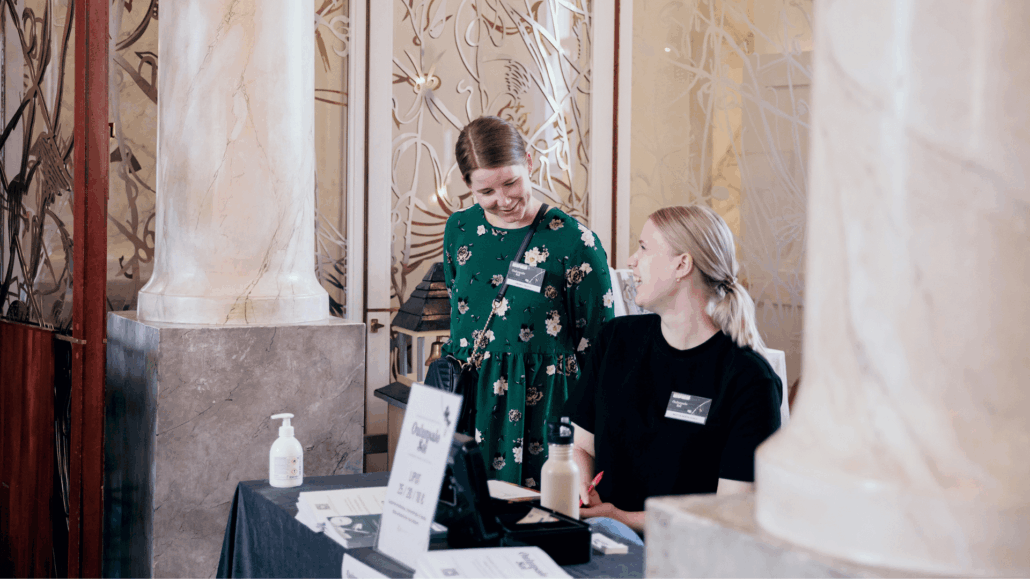 Kaksi naishenkilöä hymyilevät toisilleen lipunmyyntitiskin takana. Toisella on yllään vihreä mekko ja toisella musta paita.