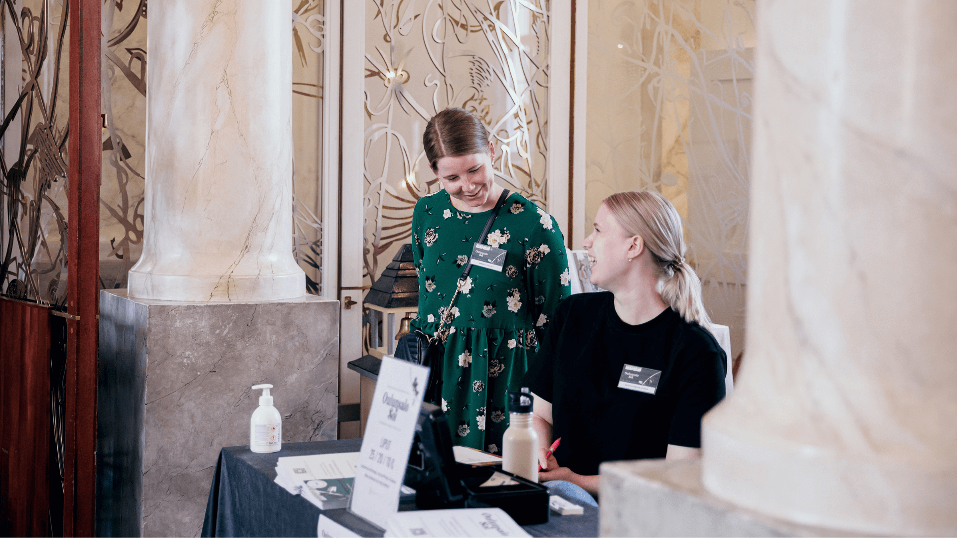 Kaksi naishenkilöä hymyilevät toisilleen lipunmyyntitiskin takana. Toisella on yllään vihreä mekko ja toisella musta paita.