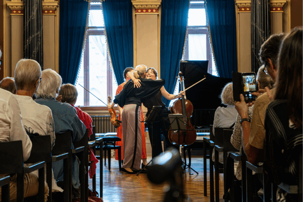 Kaksi muusikkoa halaa toisiaan hymyilleen ja yleisö istuu oikealla ja vasemmalla laidalla.