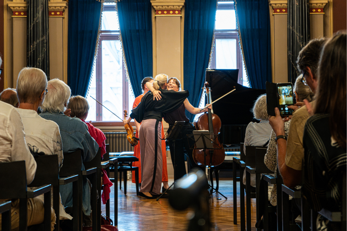 Kaksi muusikkoa halaa toisiaan hymyilleen ja yleisö istuu oikealla ja vasemmalla laidalla.