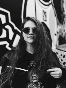 mustavalkoisessa kuvassa pitkähiuksinen nainen mustissa vaatteissa ja aurinkolaseissa, taustalla seinämaalaus