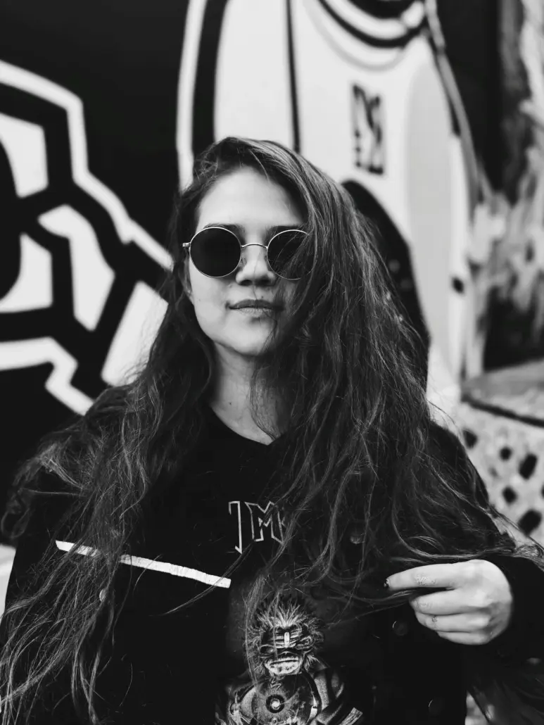 mustavalkoisessa kuvassa pitkähiuksinen nainen mustissa vaatteissa ja aurinkolaseissa, taustalla seinämaalaus
