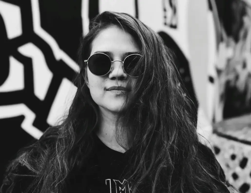 mustavalkoisessa kuvassa pitkähiuksinen nainen mustissa vaatteissa ja aurinkolaseissa, taustalla seinämaalaus