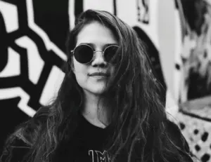 mustavalkoisessa kuvassa pitkähiuksinen nainen mustissa vaatteissa ja aurinkolaseissa, taustalla seinämaalaus