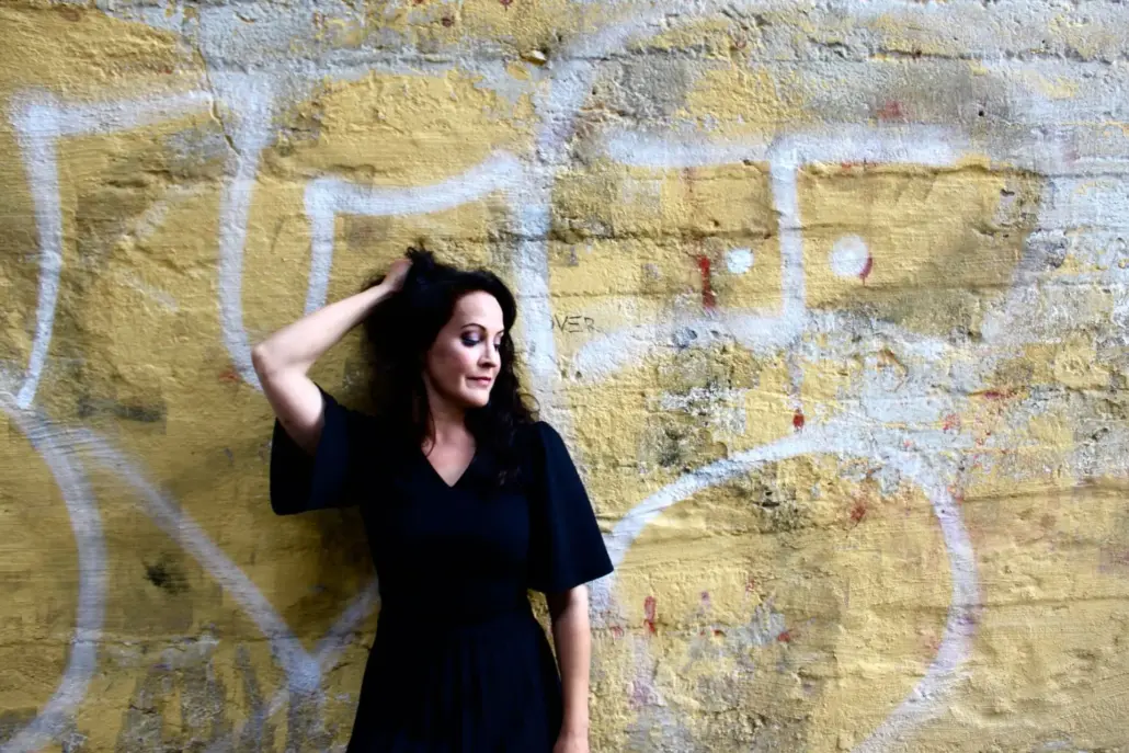 nainen mustassa mekossa pitää toista kättä pään takana ja katsoo alaspäin, taustalla vaalea graffitiseinä